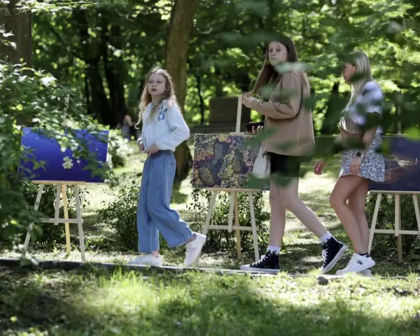 Trzy dziewczyny spacerują wśród obrazów na sztalugach. Jedna z nich, w niebieskich jeansach, przypomina młodszą wersję Tomasza Kłosowskiego.