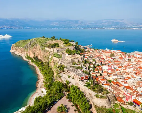 Widok na Nafplio w Grecji: miasto z czerwonymi dachami, twierdza na skale i biały statek wycieczkowy na lazurowym morzu.