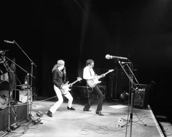 Deux guitaristes d'un groupe français rock jouent sur scène, éclairés par des projecteurs.