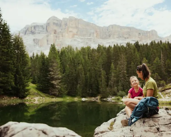 Mamma e figlia sedute su una roccia vicino a un lago alpino, con uno zaino blu. Un momento di relax per il loro lech dla lunch, con montagne imponenti sullo sfondo.