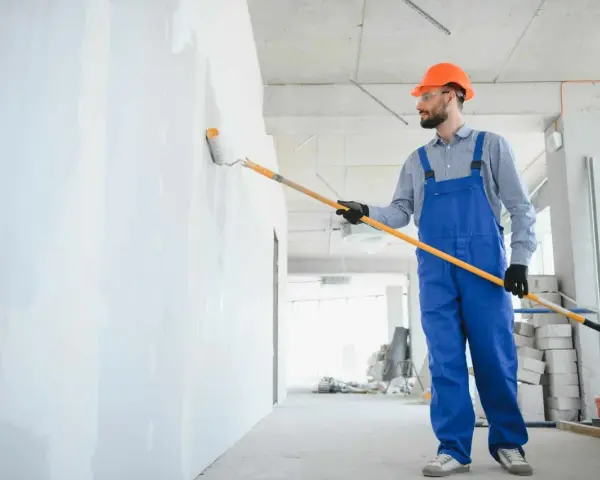 Maler mit blauer Latzhose und orangefarbenem Helm streicht eine Wand weiß. Informieren Sie sich über die malerarbeiten kosten für Ihr Projekt.