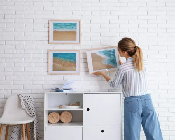 Mujer cuelga cuadros de paisajes marinos en pared de ladrillo blanco. Aprende como colocar cuadros de diferentes tamaños para un diseño armonioso.