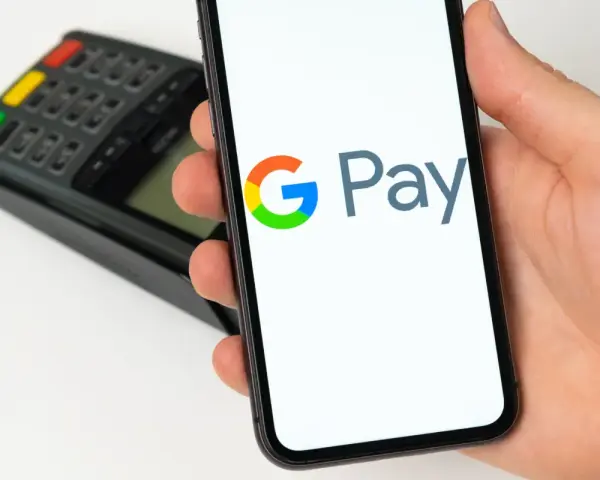 Dłoń trzyma smartfon z widocznym logo Google Pay. W tle terminal płatniczy. Szybkie i wygodne płatności.