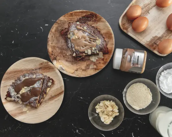 Pyszny keto omlet na słodko z kremem migdałowym, płatkami migdałów i cukrem pudrem. Obok składniki: jajka, mleko, mąka migdałowa, erytrytol.