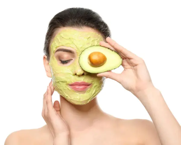 Frau mit Avocado-Gesichtsmaske und Avocado-Scheibe vor dem Auge. Die Avocado-Wirkung auf die Haut ist bekannt für ihre pflegenden Eigenschaften.