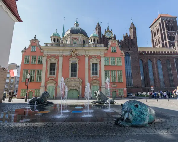 Fontanna Czterech Kwartałów w Gdańsku z lwami i wodnymi strumieniami przed różową kamienicą i bazyliką.