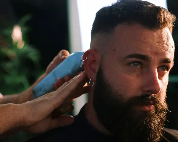 Un hombre con barba y aros en las orejas recibe un corte de pelo moderno, anticipando las tendencias de corte de pelo hombre 2026.