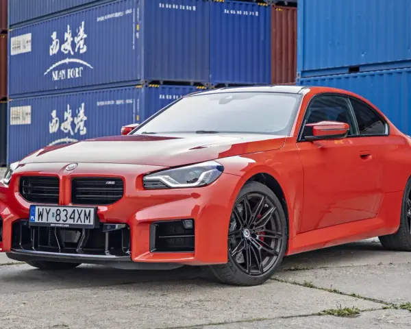 Pomarańczowe BMW M2 z czarnymi felgami, na tle niebieskich kontenerów. Sprawdź, jaka jest aktualna bmw m2 cena.