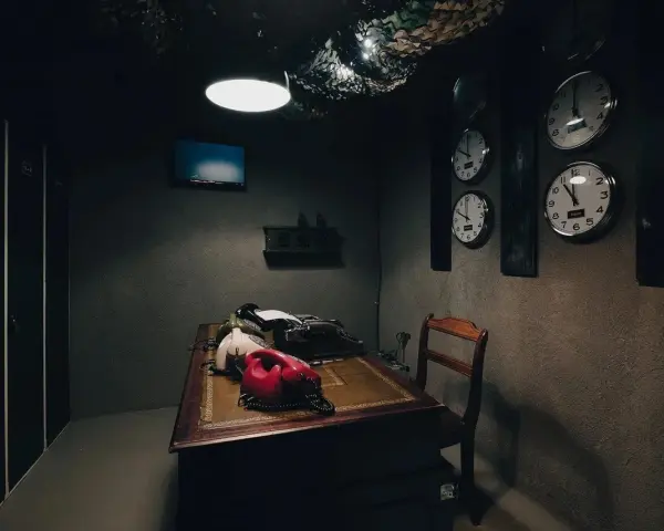 Mörkt rum med skrivbord, gamla telefoner, skrivmaskin och flera klockor på väggen. Perfekt för en spännande escape room hemma-upplevelse.