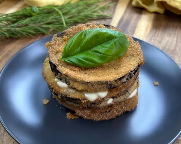Deliziose cotolette di melanzane impilate, farcite con formaggio cremoso e guarnite con una foglia di basilico fresco.