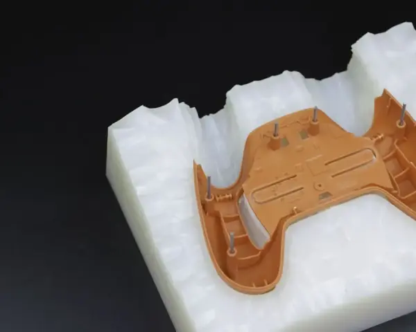 A brown plastic part, likely from a game controller, is placed on a white, textured material. This setup suggests a step in the polyurethane vacuum casting process, where a mold is being prepared.