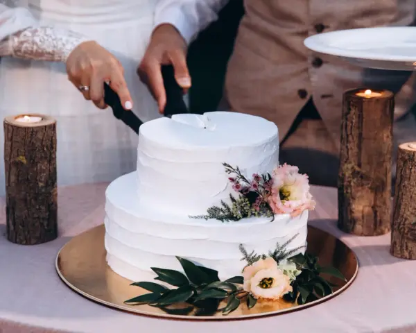 Biały tort weselny z teksturą, ozdobiony kwiatami. Para kroi tort, a obok płoną świece w drewnianych świecznikach. Trendy w prezentacji tortów weselnych to m.in. tekstury.
