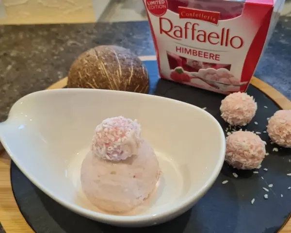 Ein Löffel Raffaello Himbeer Eis in einer Schale, daneben drei Raffaello-Kugeln und eine Kokosnuss.