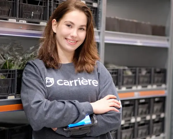 Young woman in a grey sweatshirt with "carrière" logo, holding a scanner, in a warehouse with shelves of black crates.