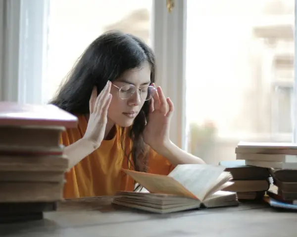 Ragazza con occhiali e mani sulle tempie, circondata da libri. Potrebbe soffrire di mancanza di concentrazione cause.