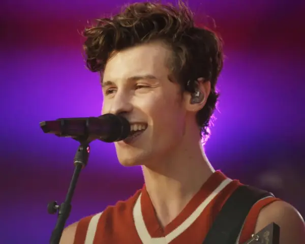 Shawn Mendes śpiewa swoje utwory na koncercie, z mikrofonem przed ustami i gitarą na ramieniu.