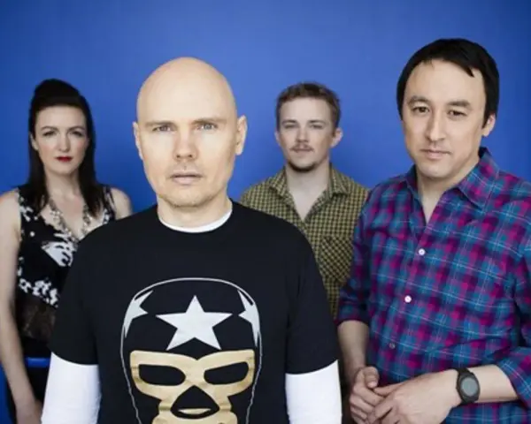 Les membres de The Smashing Pumpkins posent devant un fond bleu. Billy Corgan porte un t-shirt noir avec un masque de lucha.