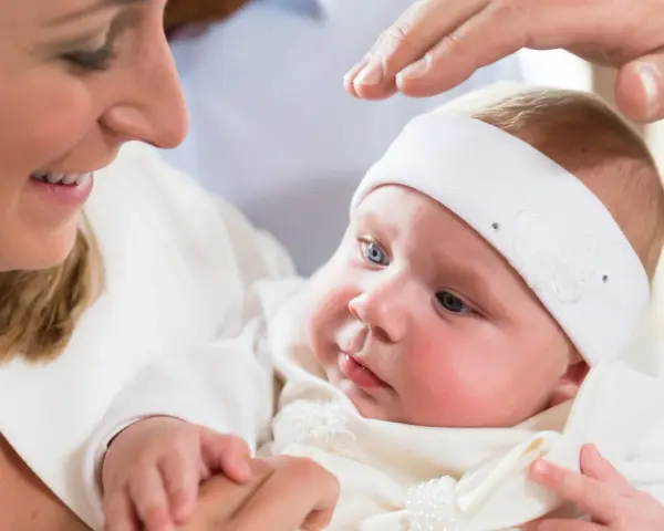 Uśmiechnięta mama trzyma dziecko w białej szatce. Dłoń ojca błogosławi malucha. Zastanawiasz się, ile kosztuje chrzest?