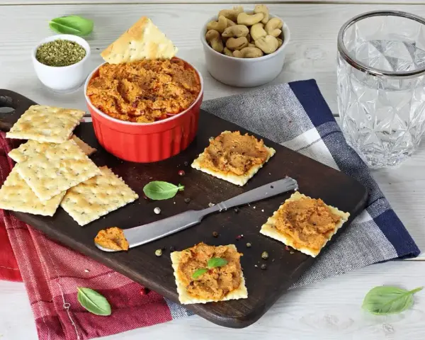 Pyszna pasta z suszonych pomidorów na krakersach, obok miseczka orzechów i szklanka wody. Idealna przekąska!