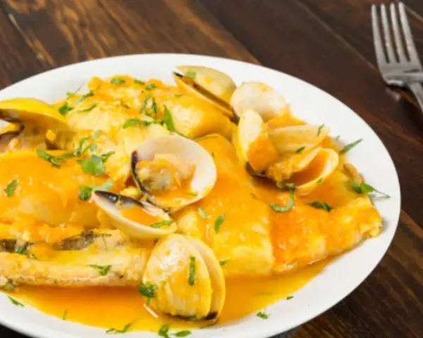 Merluza a la marinera con almejas, servida en un plato blanco sobre una mesa de madera oscura.