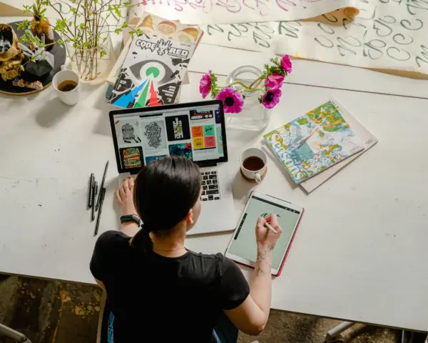 Kobieta pracuje online w domu, projektując na laptopie i tablecie. Na biurku kwiaty, kubek kawy i czaszki z pisakami.