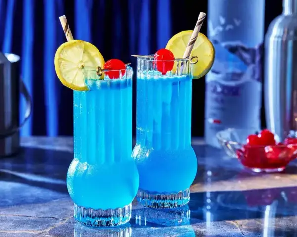 Dwa orzeźwiające, **blue drinki** z cytryną i wiśnią, gotowe do podania. W tle widać butelkę wódki i shaker.