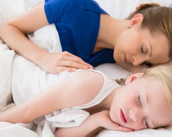 Mama i dziecko śpią razem, otuleni białymi pościelami. Czy dzieci powinny spać z rodzicami? Ta chwila spokoju sugeruje, że tak.