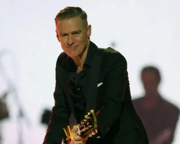Bryan Adams w czarnym garniturze gra na gitarze. Sprawdź jego nadchodzące wydarzenia i koncerty.
