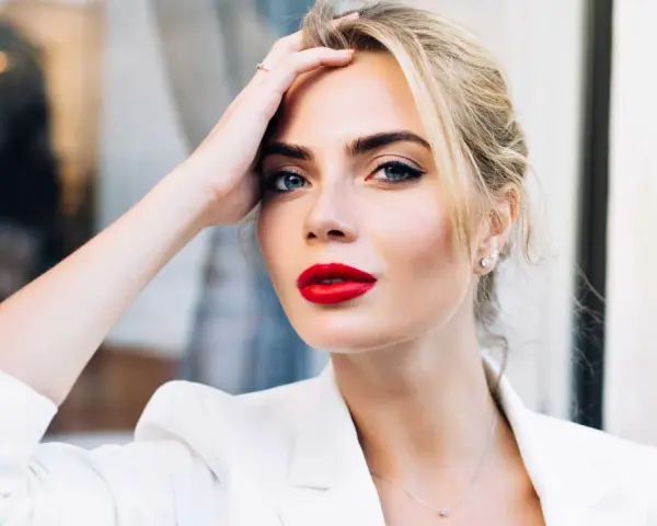 Frau mit roten Lippen und weißem Blazer, Hand am Kopf.