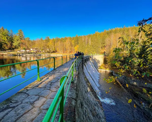 Kamienna zapora z zieloną balustradą, wodospadem i jesiennym lasem w tle.
