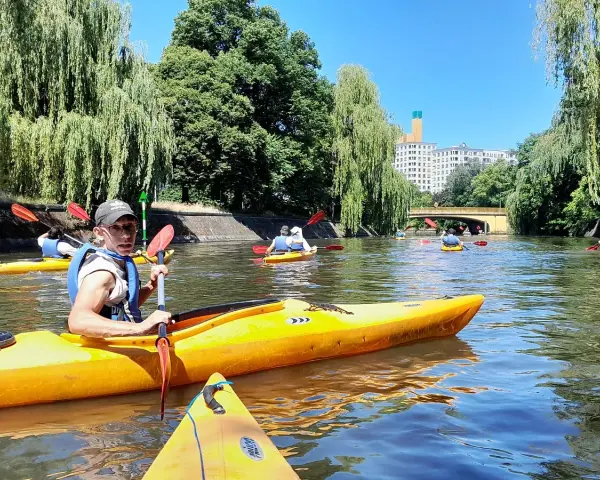 Eine Gruppe von Menschen paddelt auf gelben Kajaks auf einem Fluss in Berlin. Bäume säumen das Ufer, im Hintergrund ist ein Gebäude zu sehen.