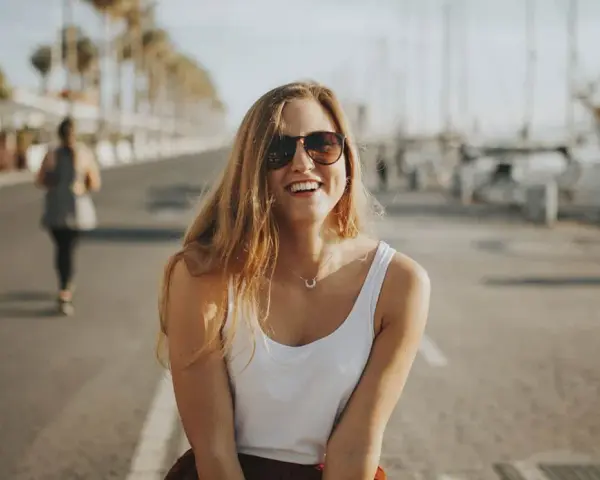 Glückliche junge Frau mit Sonnenbrille und weißem Top genießt ihr Praktikum in Spanien am Hafen.