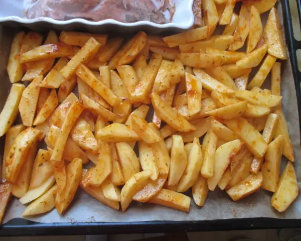 Gotowe do pieczenia frytki w piekarniku, pokrojone w łódeczki i doprawione przyprawami, leżą na blasze wyłożonej papierem.