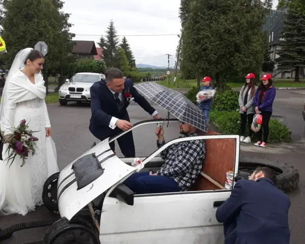 Brama weselna z udziałem strażaka, pana młodego i panny młodej. Pan młody rozmawia z druhem siedzącym w prowizorycznym samochodzie.