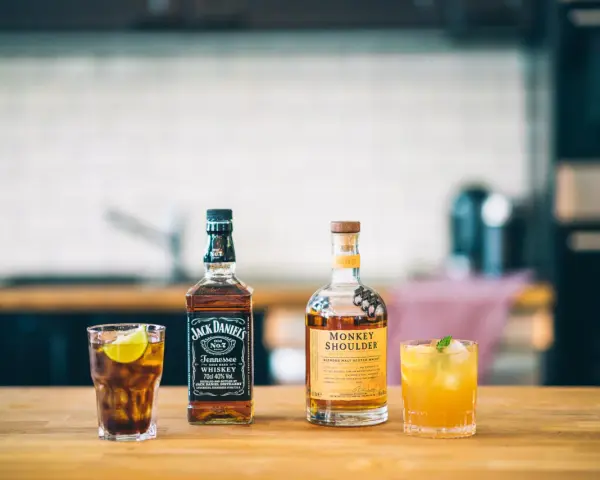 Zwei Flaschen Whiskey, Jack Daniel's und Monkey Shoulder, stehen neben zwei Cocktails auf einem Holztisch.