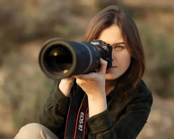 Kobieta z aparatem Canon EOS R7, skupiona na kadrze. Jej oczy błyszczą, a obiektyw sugeruje zaawansowane techniki fotograficzne.