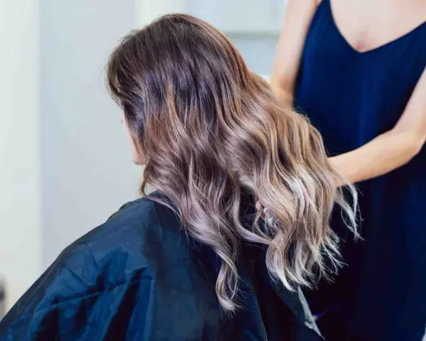 Kobieta z falowanymi włosami w odcieniach brązu i subtelnym balejażem blond. Fryzjerka poprawia jej fryzurę.