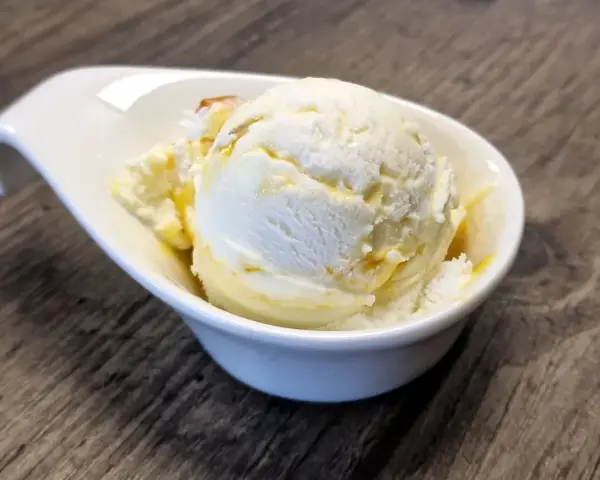 Ein Löffel cremiges Buttermilch-Eis, beträufelt mit Honig, in einer weißen Schale auf Holztisch.