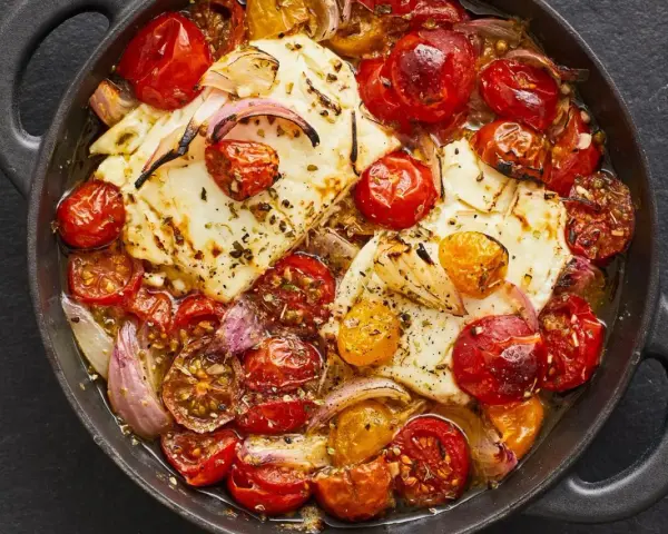 Heißer, gebackener Feta mit Kirschtomaten und roten Zwiebeln in einer Pfanne, mit Kräutern bestreut.