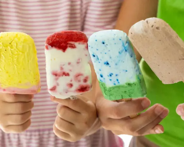 Vier Kinderhände halten bunte Eis am Stiel. Ein gelbes, ein Erdbeer-Vanille, ein blau-grünes und ein Schokoladen-Eis. Perfekt für heiße Tage!