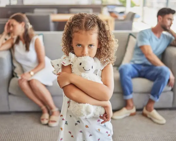 Ein Kind umklammert sein Stofftier, während die Eltern im Hintergrund getrennt sitzen. Ein Bild, das die emotionale Belastung eines Trennungsjahres mit Kindern widerspiegelt.