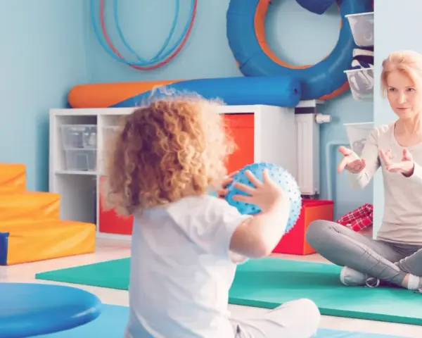 Ergotherapie für Kinder: Eine Therapeutin erklärt einem Kind mit Locken, wie man einen Ball wirft.