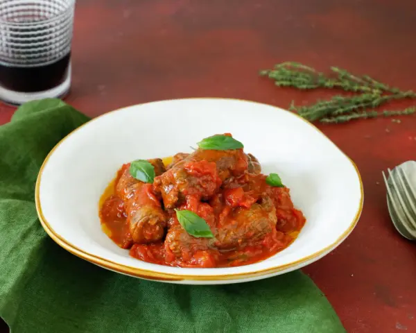 Involtini di carne in padella con sughetto, guarniti con basilico fresco, serviti in un piatto bianco su un tovagliolo verde.