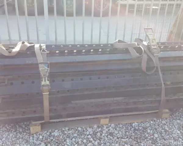 Stos czarnych, metalowych elementów naczepy z pasami transportowymi.