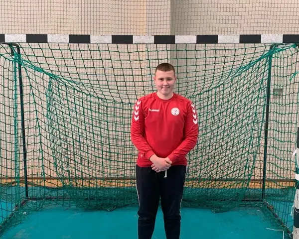 Dawid Kozak w czerwonej bluzie z długim rękawem i czarnych spodniach stoi przed bramką do piłki ręcznej.
