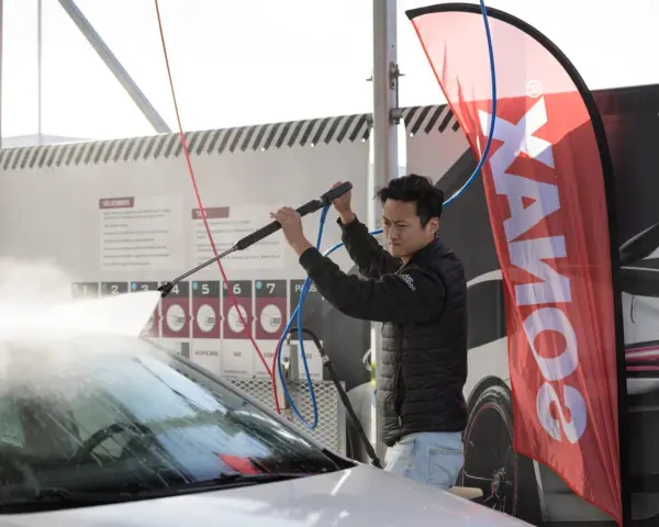 Man using a pressure washer on a car windshield, with a SONAX banner in the background.