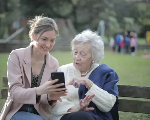 Młoda kobieta pokazuje coś na telefonie starszej pani. To scena z życia, jak z kursu opiekuna osoby starszej, gdzie uczymy się wspierać seniorów w codzienności.