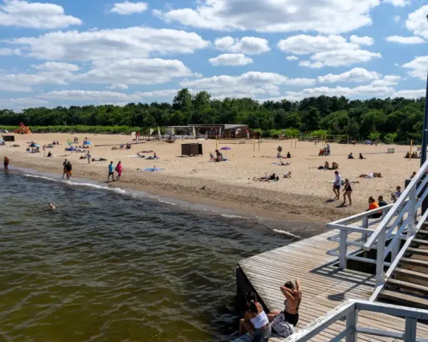 Słoneczny dzień nad morzem bałtyckim. Ludzie odpoczywają na piaszczystej plaży, niektórzy kąpią się w wodzie. Drewniane molo prowadzi na plażę.