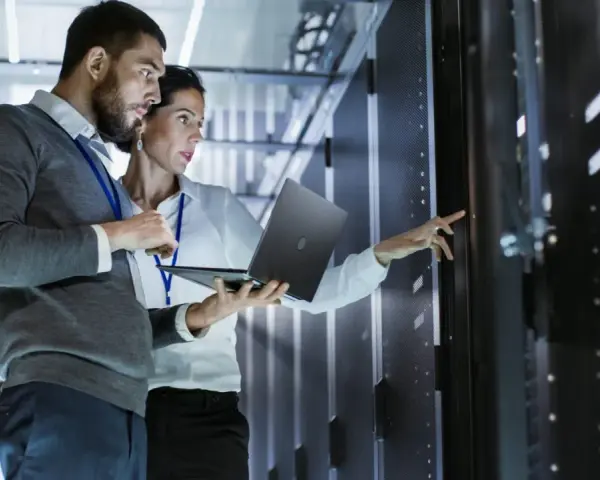 Two IT professionals collaborate on network management, one holding a laptop while the other points to a server rack.