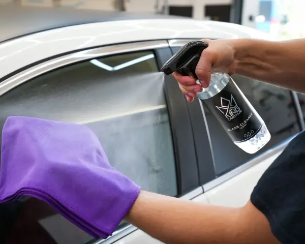 En hand sprutar rengöring på insidan av bilrutan med en mikrofiberduk redo att torka.